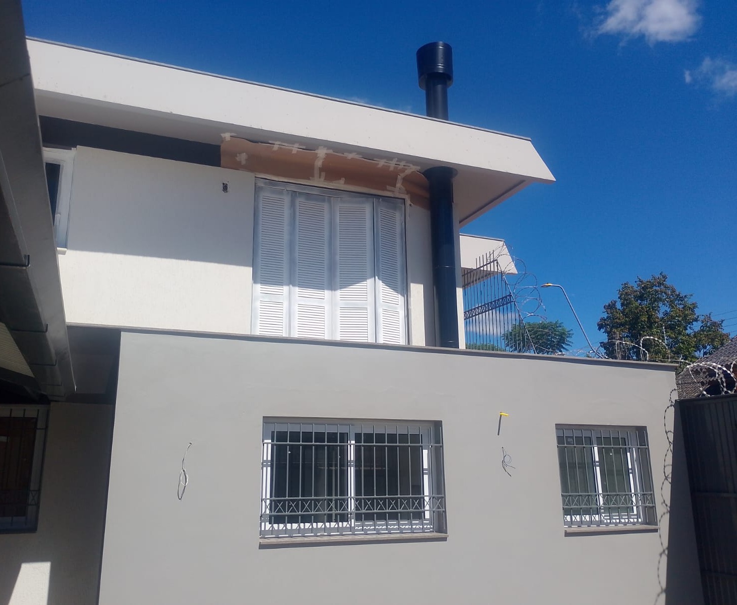 Fachada de um prédio branco com janelas gradeadas, persianas e uma chaminé preta de fogão a lenha instalada no