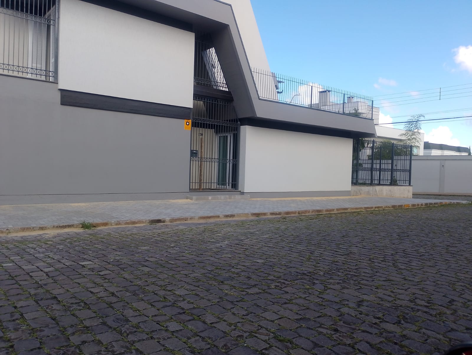 Fachada de prédio moderno em cinza e branco com portão de ferro e rua de paralelepípedos na frente